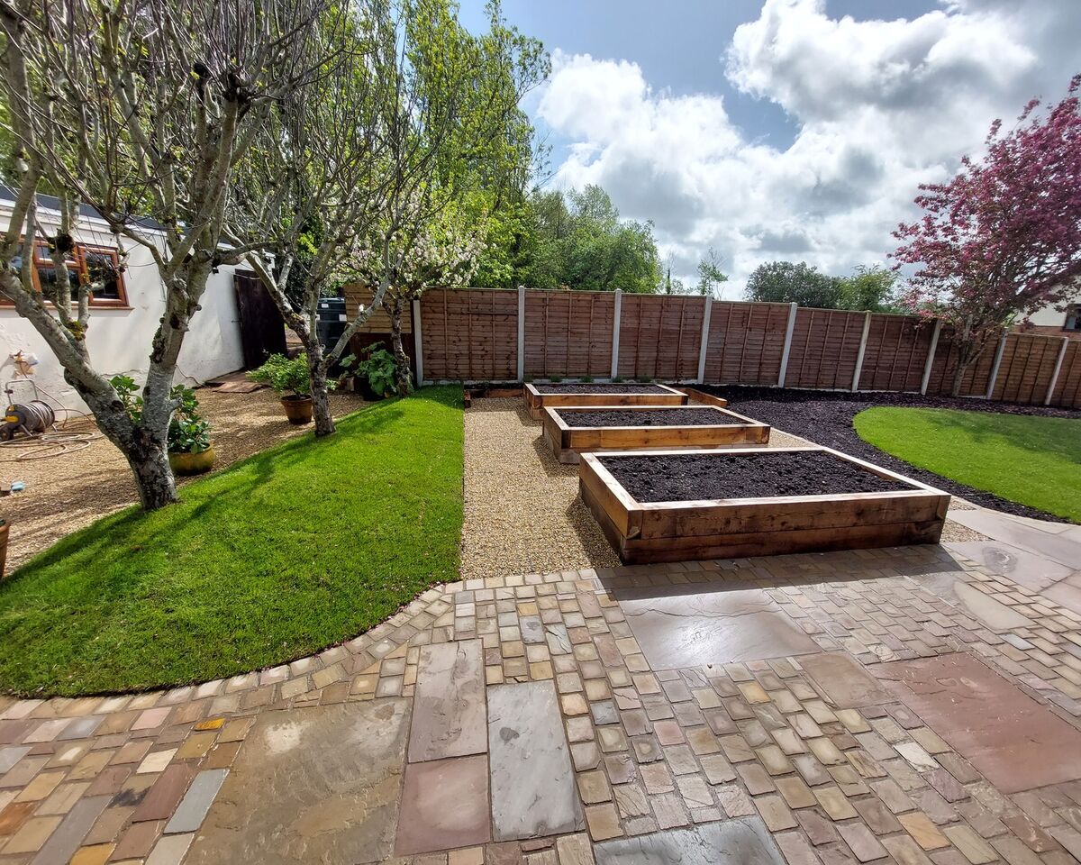 A garden with raised wooden beds, grassy areas, and a stone pathway under a cloudy sky.