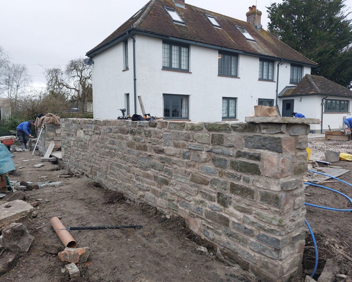 Stone wall under construction in front of a white house. Tools and materials visible nearby.