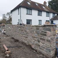 Stone wall under construction in front of a white house. Tools and materials visible nearby.
