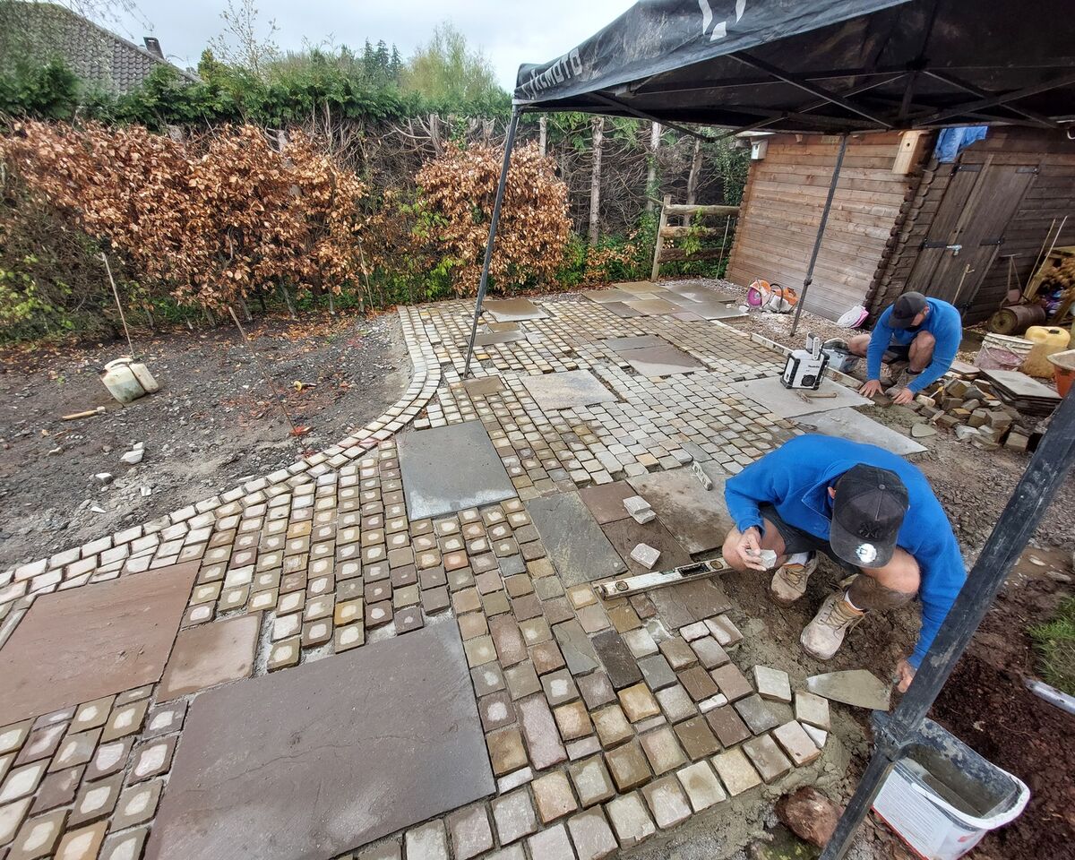 Two workers laying stones in a garden area with hedges in the background.