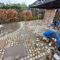 Two workers laying stones in a garden area with hedges in the background.