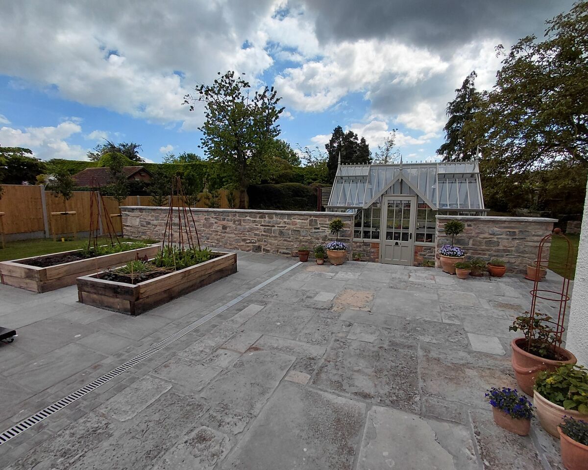 A garden patio with raised plant beds and a greenhouse in the background under cloudy skies.