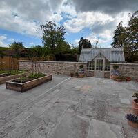A garden patio with raised plant beds and a greenhouse in the background under cloudy skies.