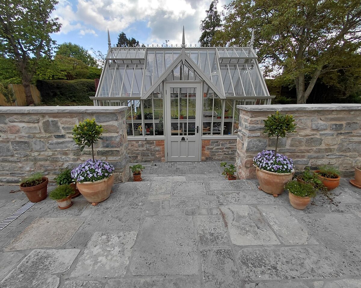 Greenhouse behind a stone wall, surrounded by potted plants and a paved pathway.