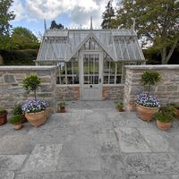 Greenhouse behind a stone wall, surrounded by potted plants and a paved pathway.