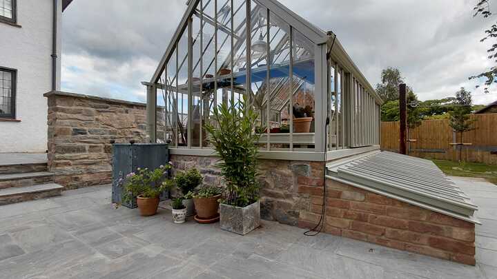 A glass greenhouse with plants, adjacent to a stone path and house, under a cloudy sky.