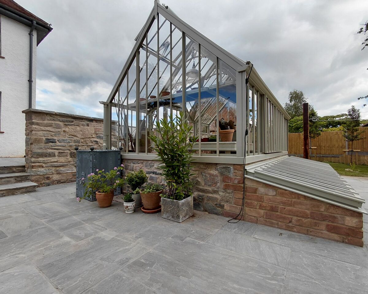 A glass greenhouse with potted plants, adjacent to a house and stone pathway.