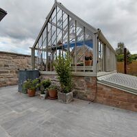 A glass greenhouse with potted plants, adjacent to a house and stone pathway.