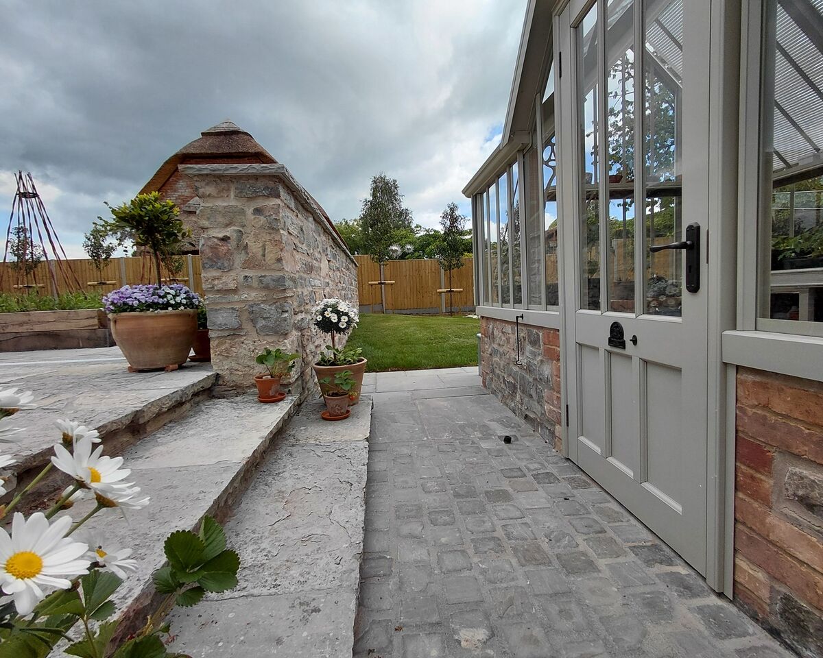 Garden setting with a stone wall, patio steps, and a greenhouse under a cloudy sky.