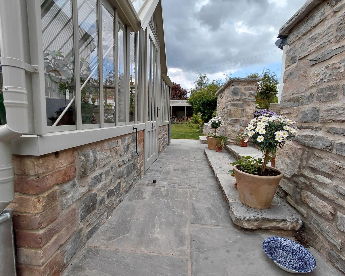 Pathway alongside a stone wall and greenhouse, with potted plants and cloudy sky.