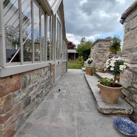 Pathway alongside a stone wall and greenhouse, with potted plants and cloudy sky.