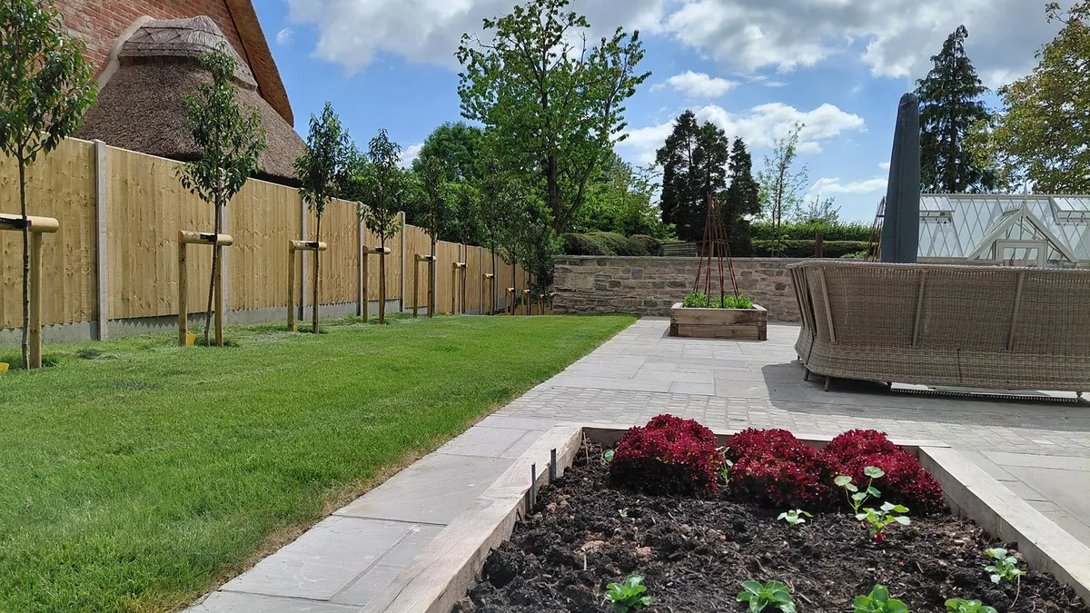 New lawn, paved terrace and raised planting beds bordered by a reclaimed stone wall, with the greenhouse beyond