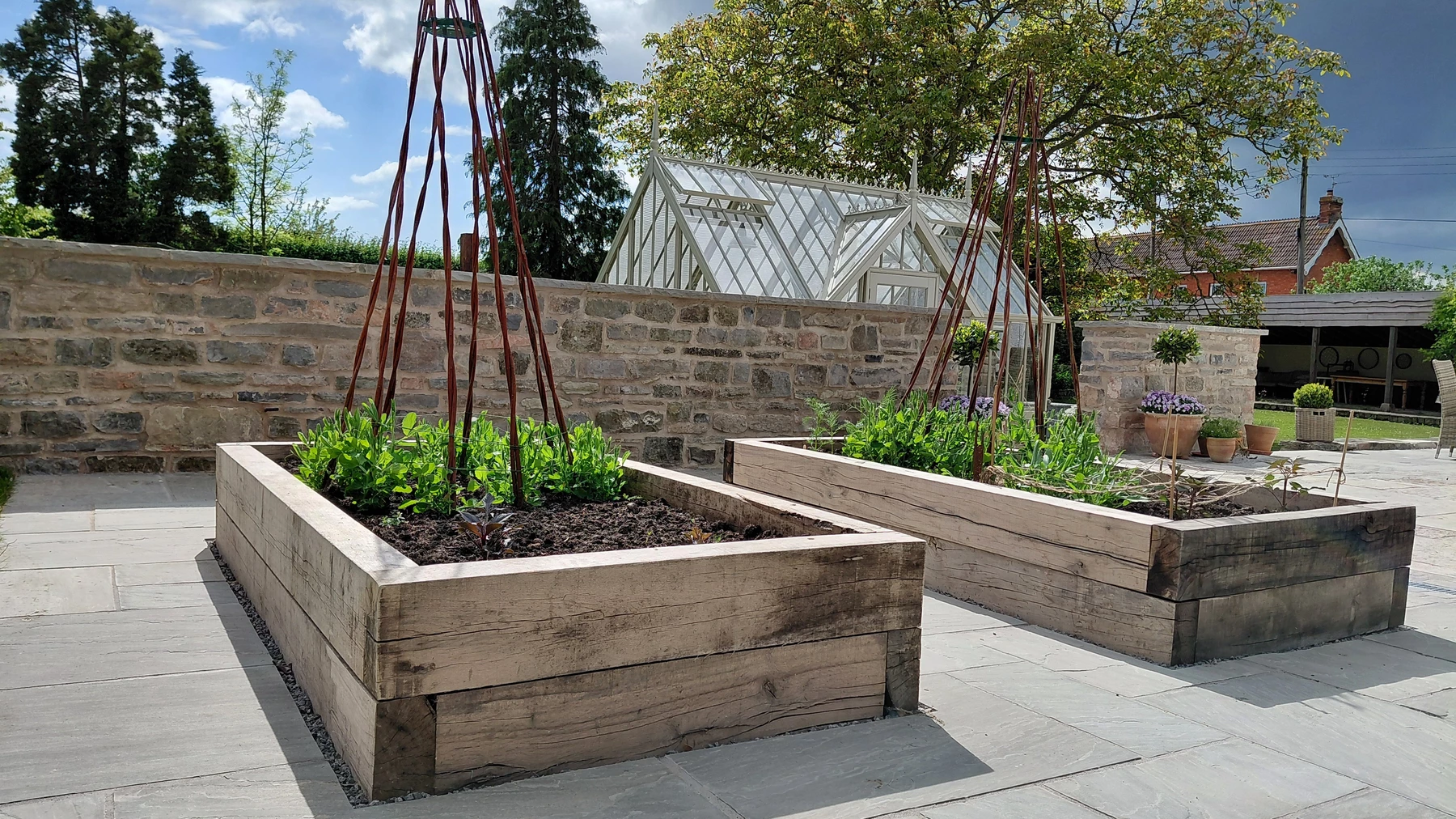 Timber raised beds with plant supports on a reclaimed stone terrace, greenhouse and stone walls in the background