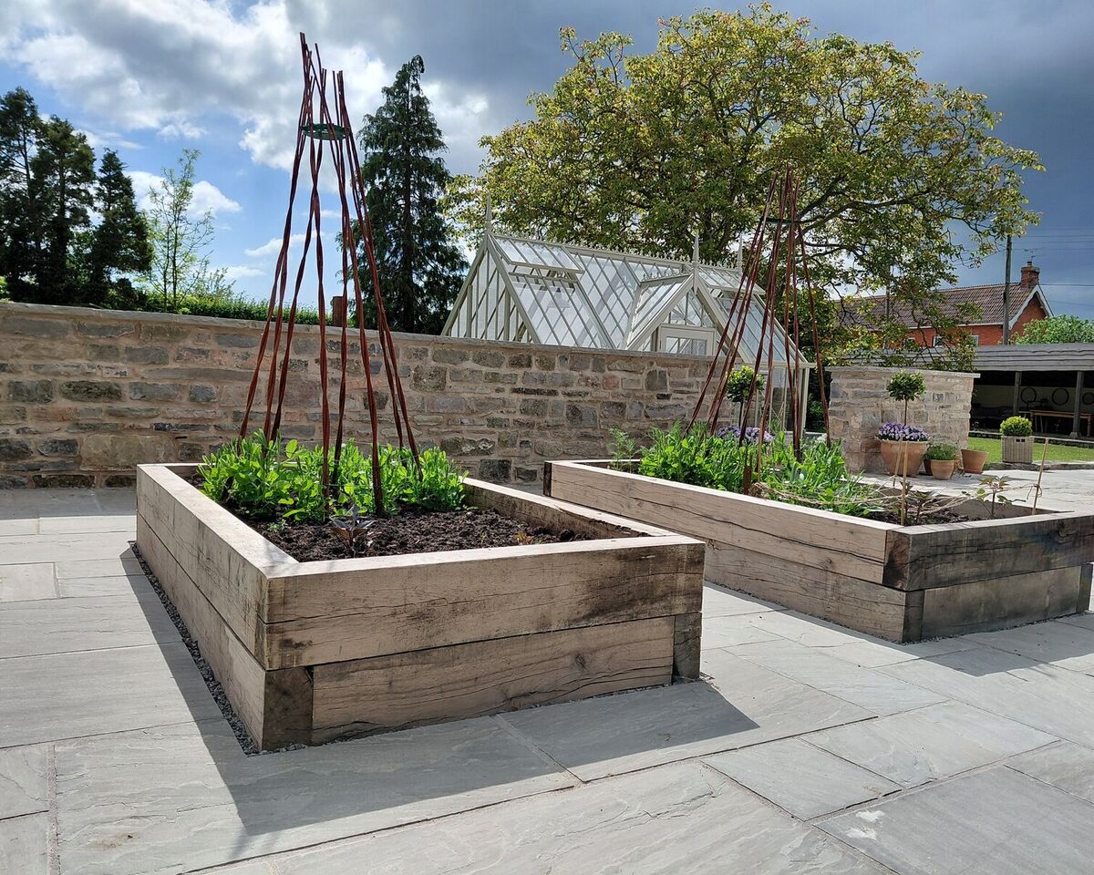 Two wooden raised garden beds with plants, surrounded by a stone wall and trees.