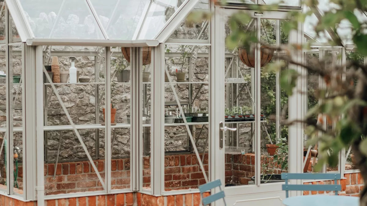 White-framed greenhouse on a brick base with potted plants, a paved path and bistro chairs against a stone garden wall