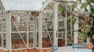 White-framed greenhouse on a brick base with potted plants, a paved path and bistro chairs against a stone garden wall