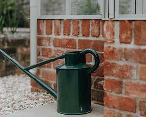 Green watering can on the pale stone paving beside the greenhouse's red brick base