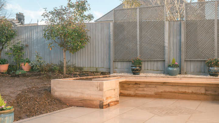 A glass greenhouse with plants, adjacent to a stone path and house, under a cloudy sky.