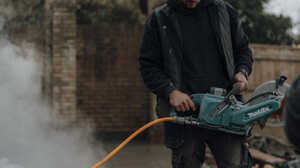 A glass greenhouse with plants, adjacent to a stone path and house, under a cloudy sky.