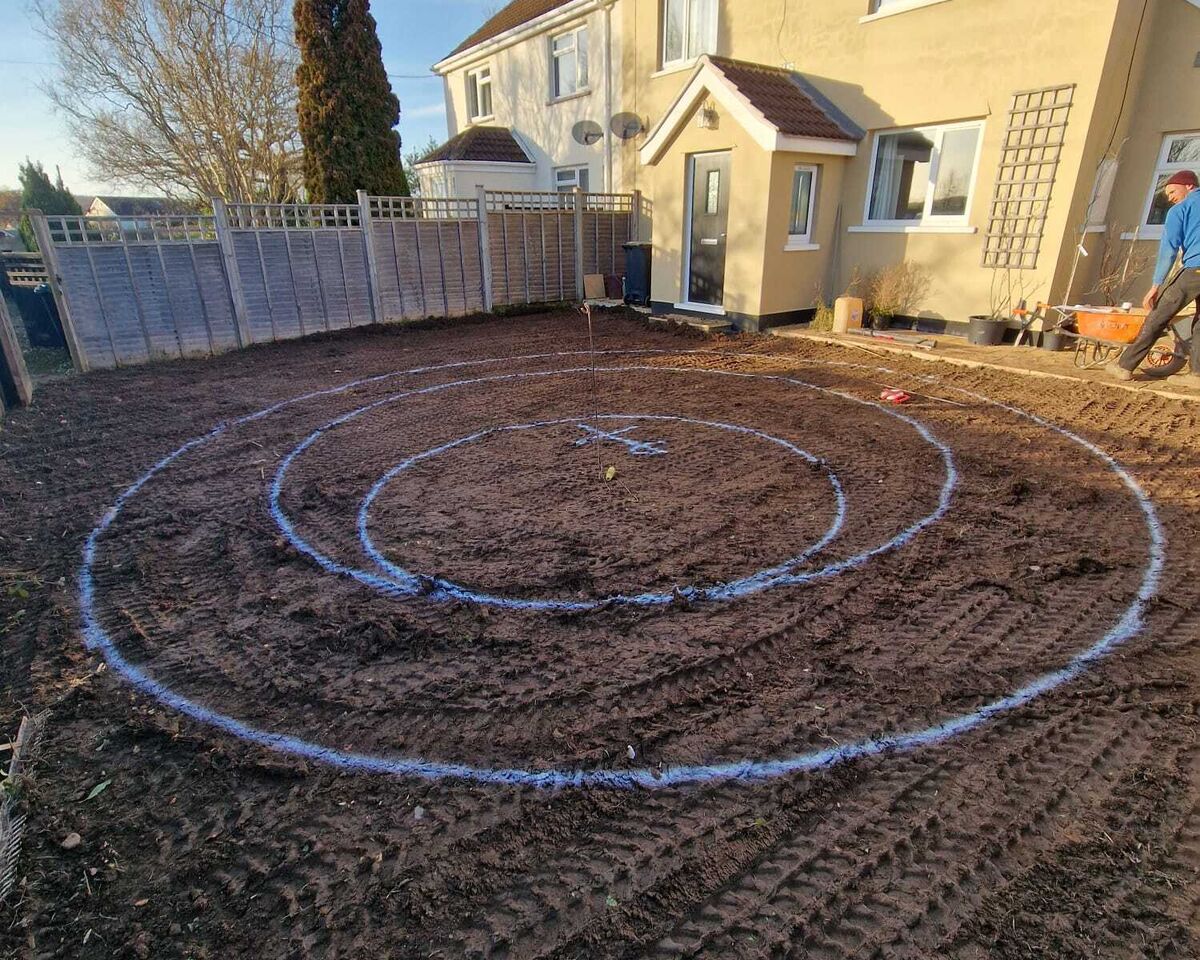 A garden with circular patterns marked out in freshly turned soil. A house is in the background.