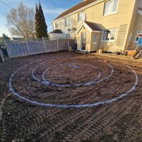 A garden with circular patterns marked out in freshly turned soil. A house is in the background.