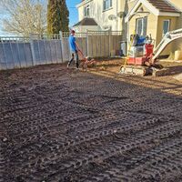 Sibley team using a tiller in a freshly tilled garden area beside a house.