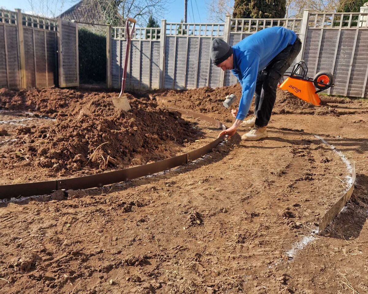 Sibley worker shapes soil in a garden, with tools and a fence in the background.