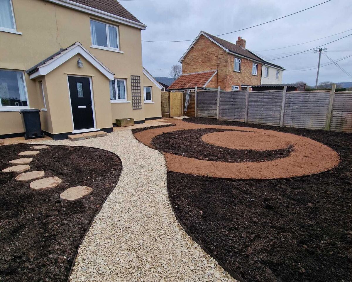 A landscaped garden with a gravel path and circular mulch design in front of two houses.