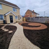 A landscaped garden with a gravel path and circular mulch design in front of two houses.