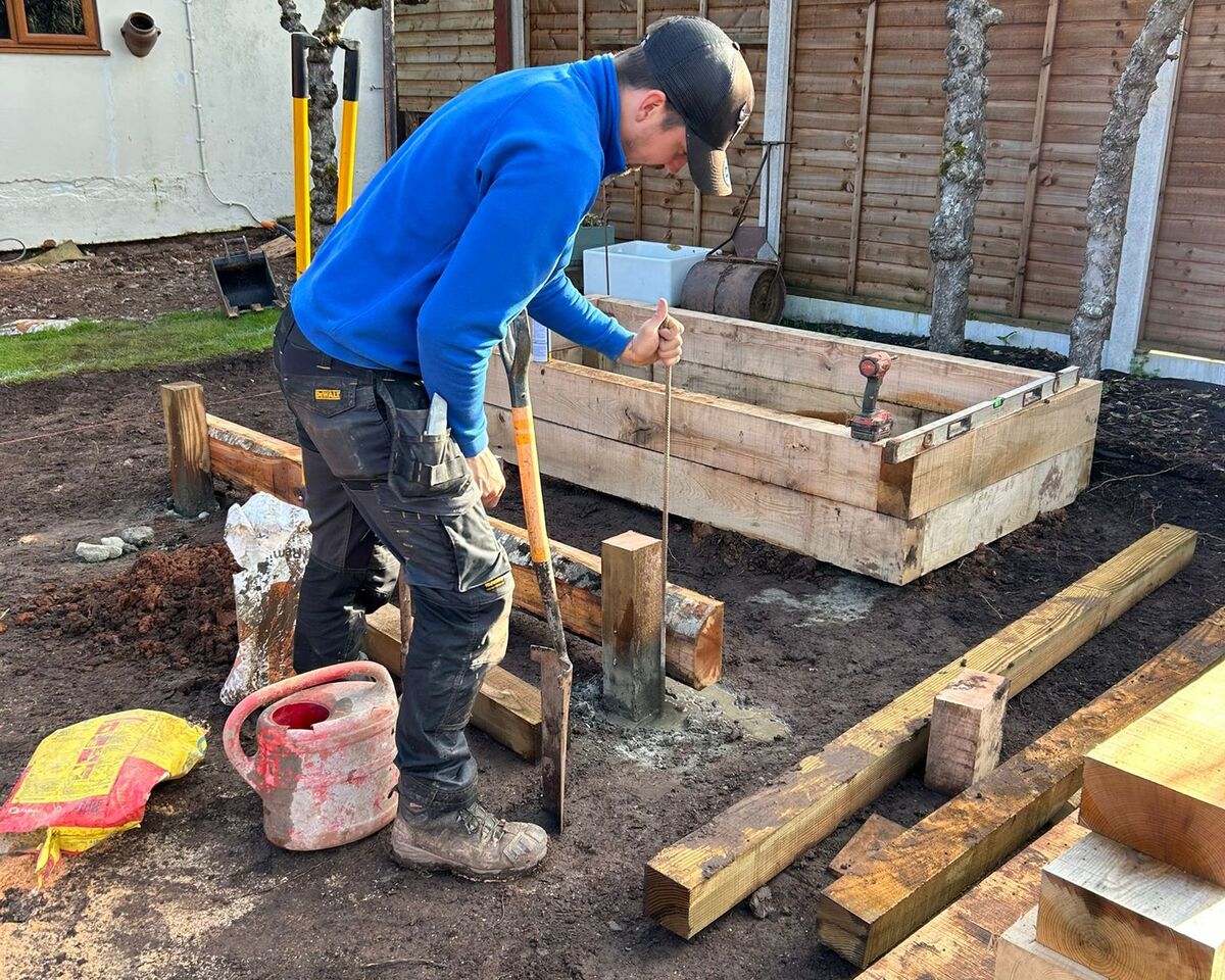 A person in a blue sweater works on a wooden structure in a garden.