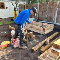 A person in a blue sweater works on a wooden structure in a garden.