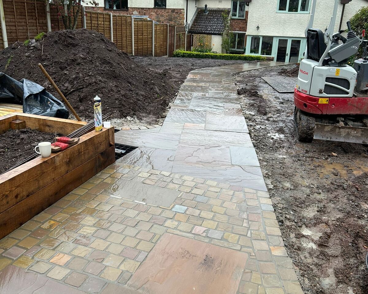 Paved pathway leading to a house, with construction materials and a machine in the background.