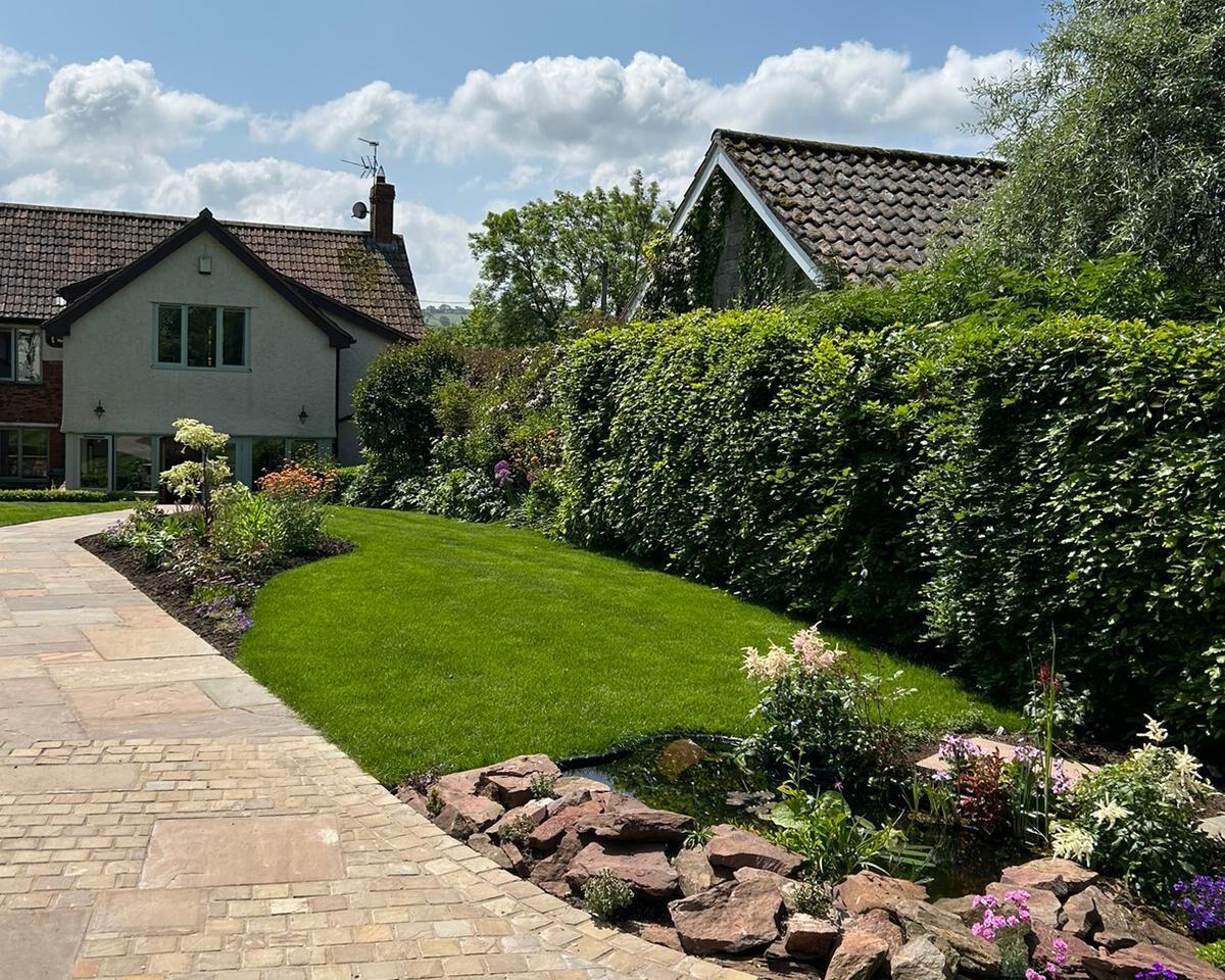 Lush garden path lined with flowers and greenery under a blue sky with clouds.