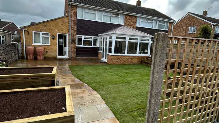 A glass greenhouse with plants, adjacent to a stone path and house, under a cloudy sky.