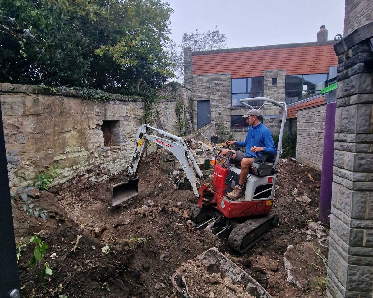 A worker operates a small excavator in a dirt-filled construction site.