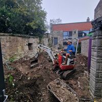 A worker operates a small excavator in a dirt-filled construction site.