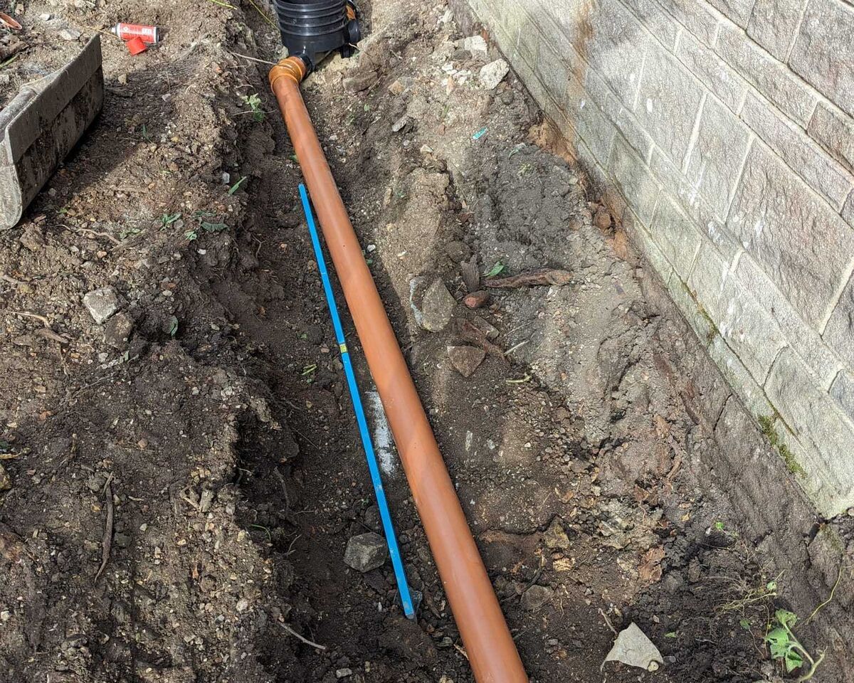 A trench with a large, orange pipe running alongside a gray wall.