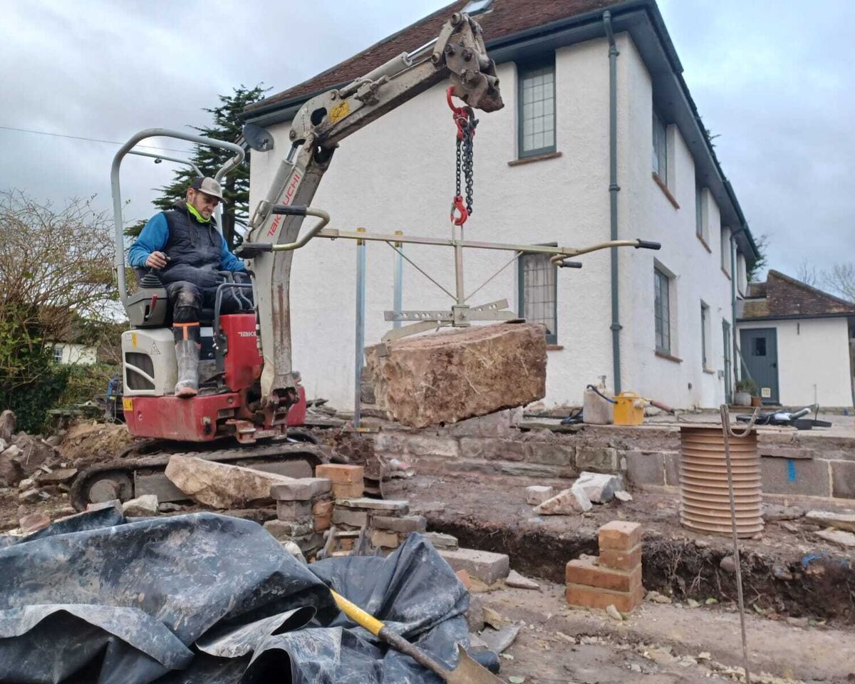 A construction worker uses machinery to lift a heavy object near a house.