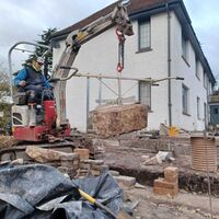 A construction worker uses machinery to lift a heavy object near a house.