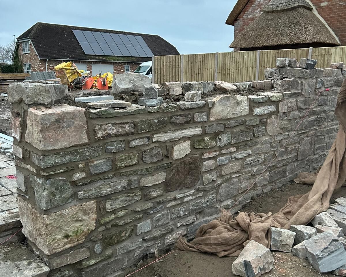 Stone wall in progress, with construction materials and a barn in the background.