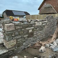 Stone wall in progress, with construction materials and a barn in the background.