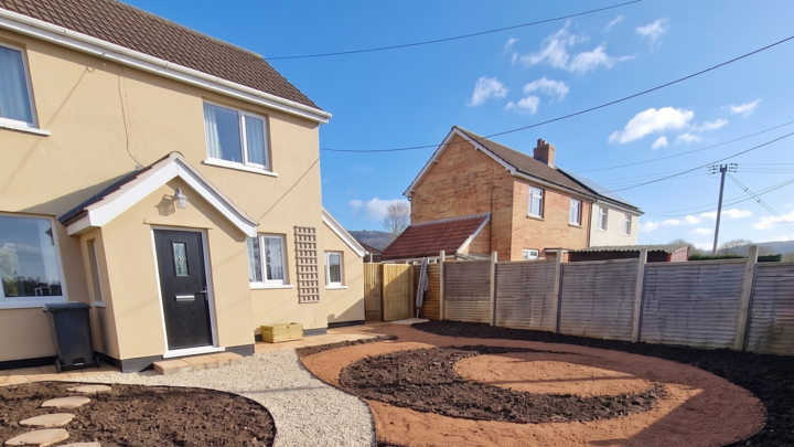 A glass greenhouse with plants, adjacent to a stone path and house, under a cloudy sky.