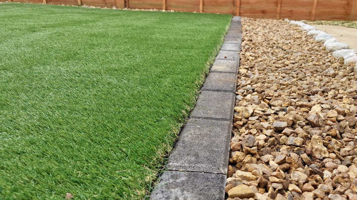 A glass greenhouse with plants, adjacent to a stone path and house, under a cloudy sky.