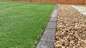 A glass greenhouse with plants, adjacent to a stone path and house, under a cloudy sky.