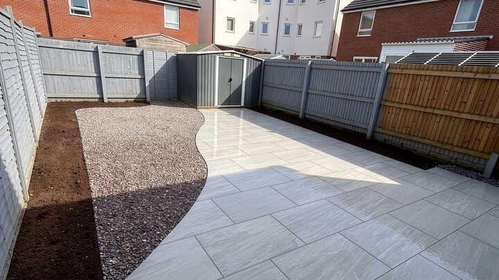 A glass greenhouse with plants, adjacent to a stone path and house, under a cloudy sky.