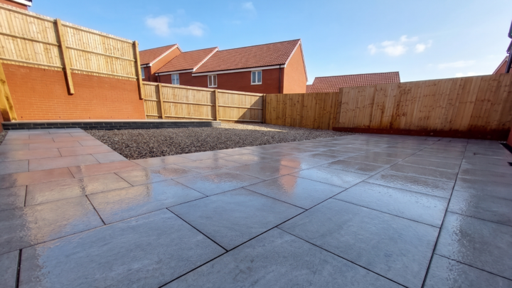 A glass greenhouse with plants, adjacent to a stone path and house, under a cloudy sky.
