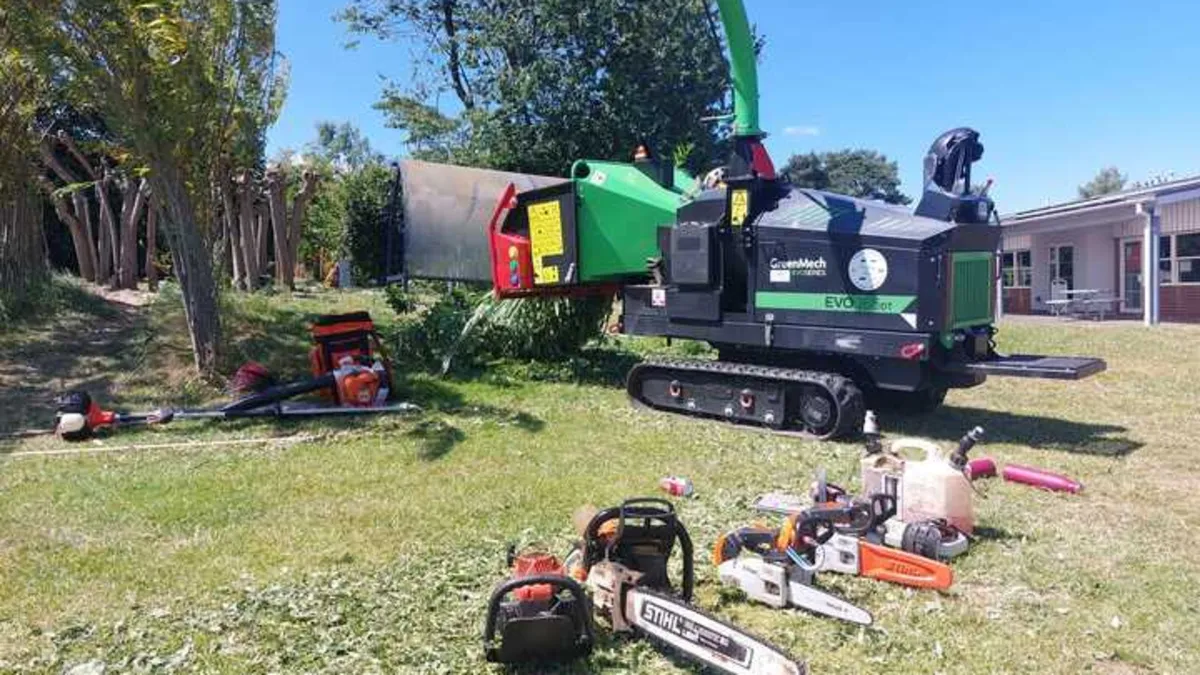 Green tracked wood chipper on a lawn with several chainsaws laid out on the grass in front