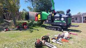 Green tracked wood chipper on a lawn with several chainsaws laid out on the grass in front